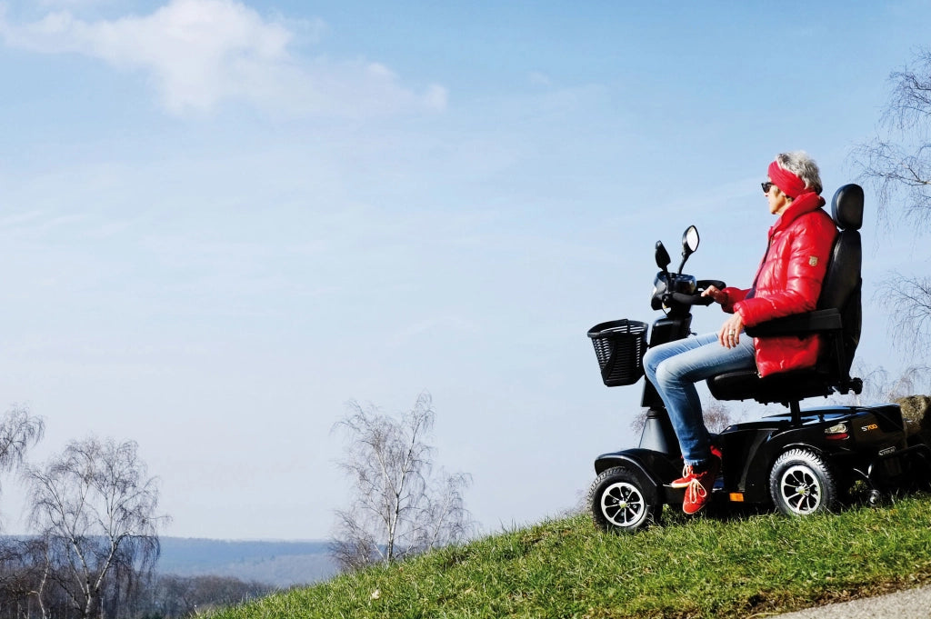 Seniorin auf einem Sterling S700 Elektromobil beim Ausflug in der Natur, fährt entspannt über eine Wiese mit weitem Ausblick – Symbol für Freiheit und aktive Mobilität im Alter.