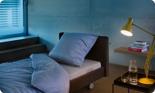 Dunkelbraunes Pflegebett mit blauem Abiente in Abendlicht, mit gelber angeschalteter Lampe