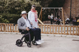 Älterer Mann mit Hut und Sonnenbrille sitzt entspannt im Rollz Motion Electric Elektrorollstuhl, während eine Frau neben ihm geht. Die Szene spielt sich in einem Park mit Spielplatz im Hintergrund ab, was die Alltagstauglichkeit und Flexibilität des Rollstuhls betont.